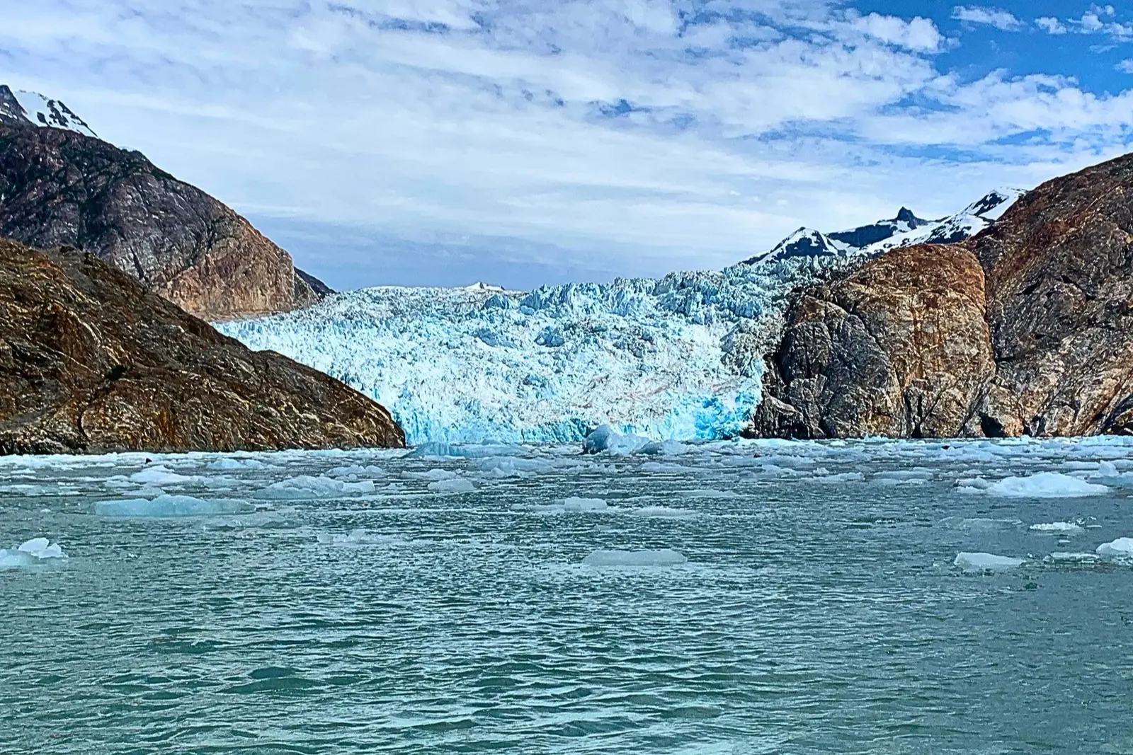 A tidewater glacier between brown rocky walls, with floating ice across the water in front.