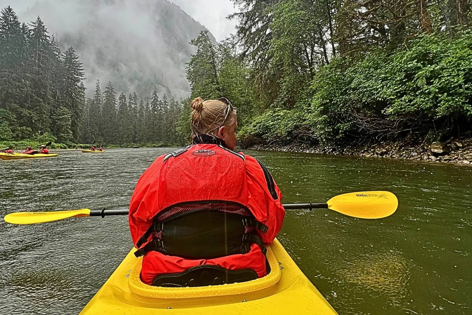 The view forward from a kayak toward a forested river mouth in the rain, with other kayaks ahead.