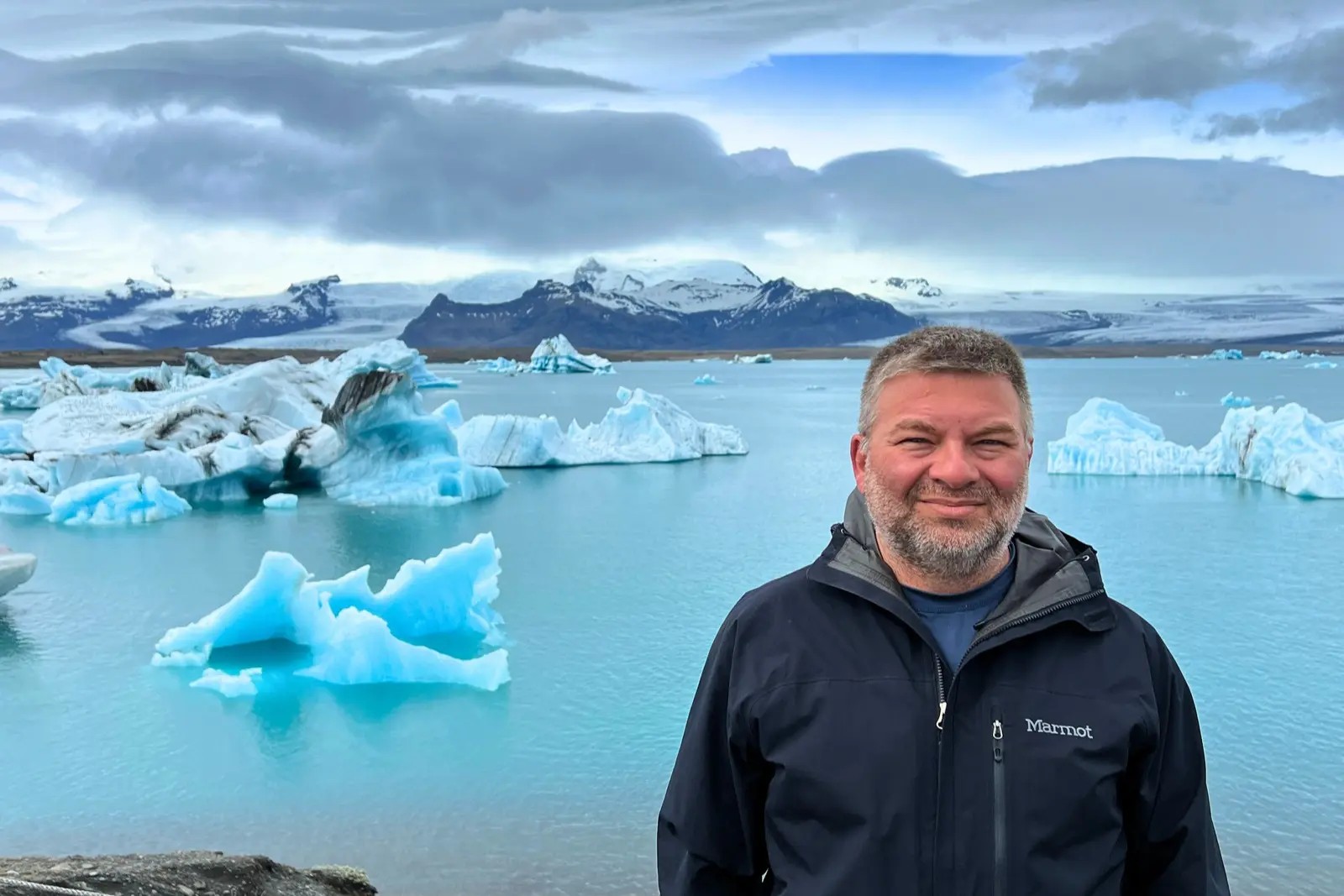 Chris Commisso in front of blue icebergs in Jökulsárlón, with a glacier and snowy mountains behind.