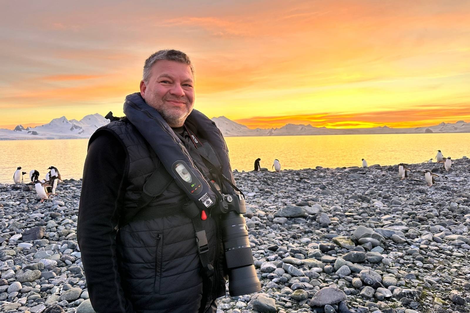 Chris Commisso standing among Gentoo penguins on the rocky shore at Yankee Harbor beneath an orange sunset.
