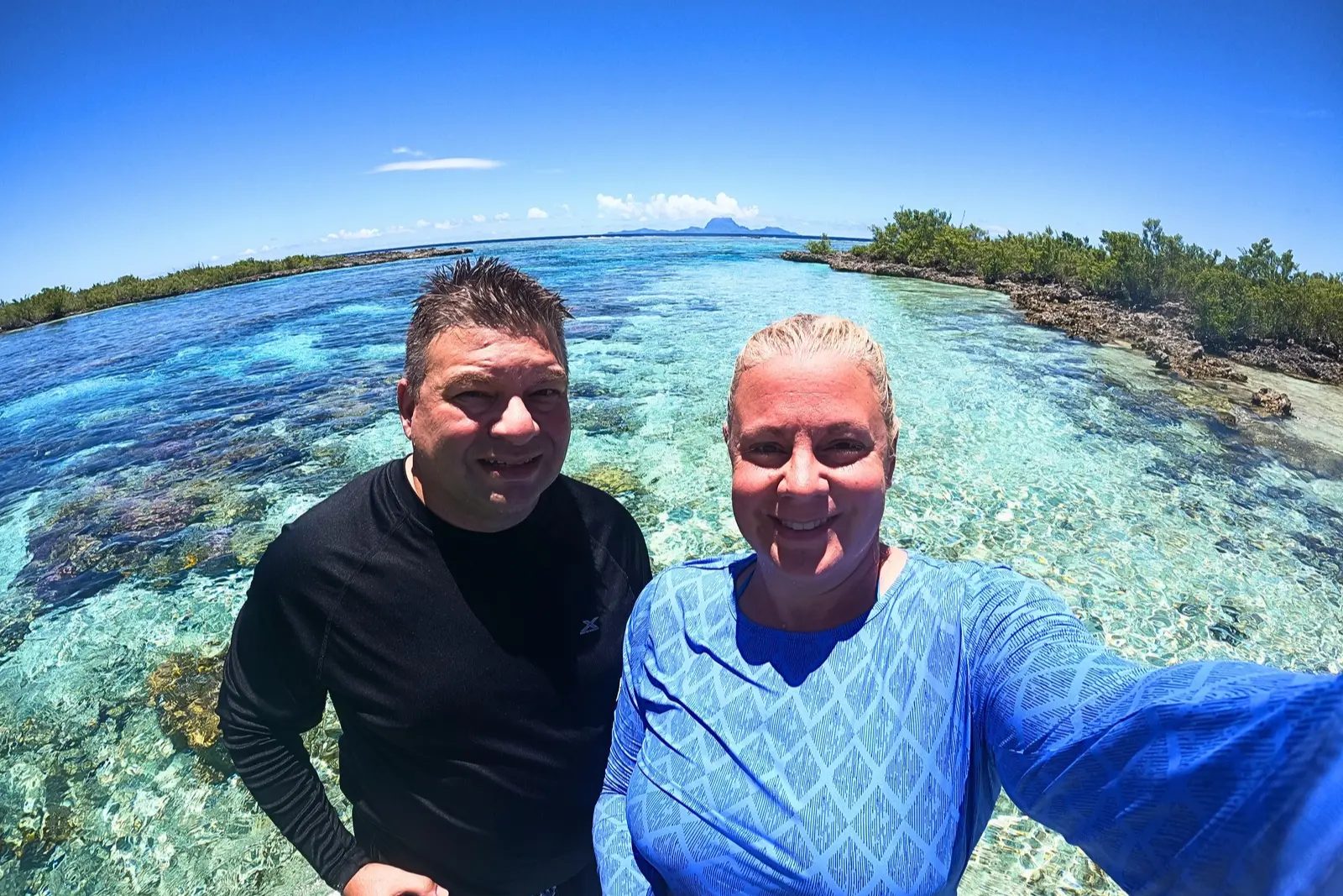 Chris and Erika Commisso standing in clear, shallow water in the drift channel at Taha'a, with Bora Bora on the horizon.