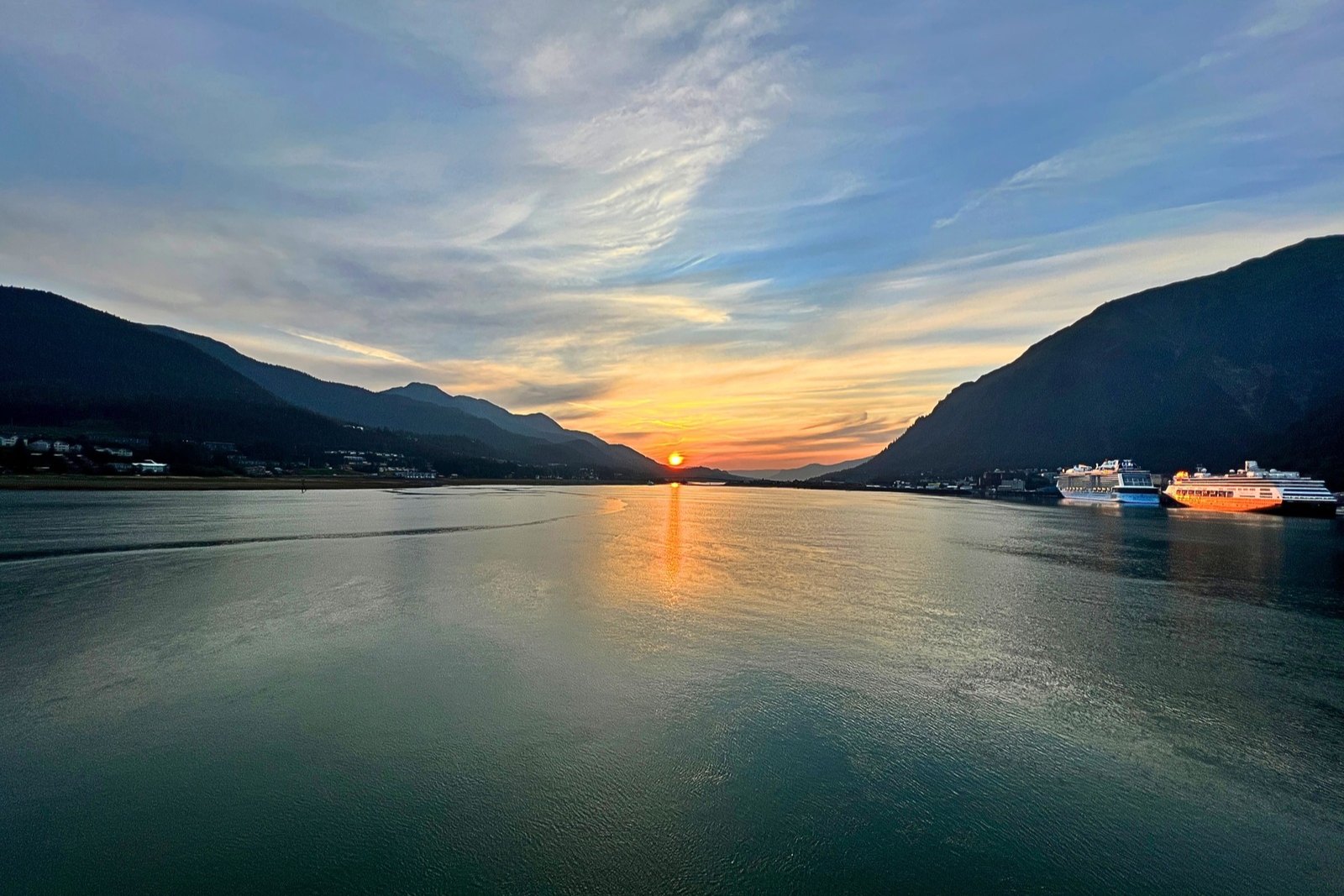 The sun setting between mountains over Gastineau Channel, with cruise ships docked along the Juneau waterfront.