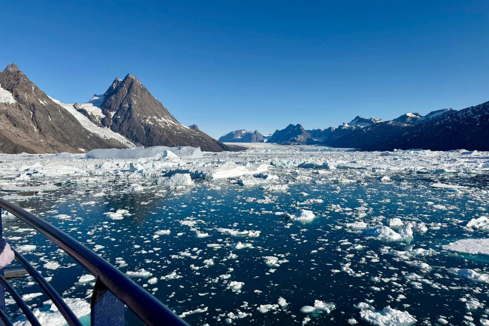 A Greenland fjord filled with floating ice, steep rocky mountains, and the railing of a ship in the foreground.