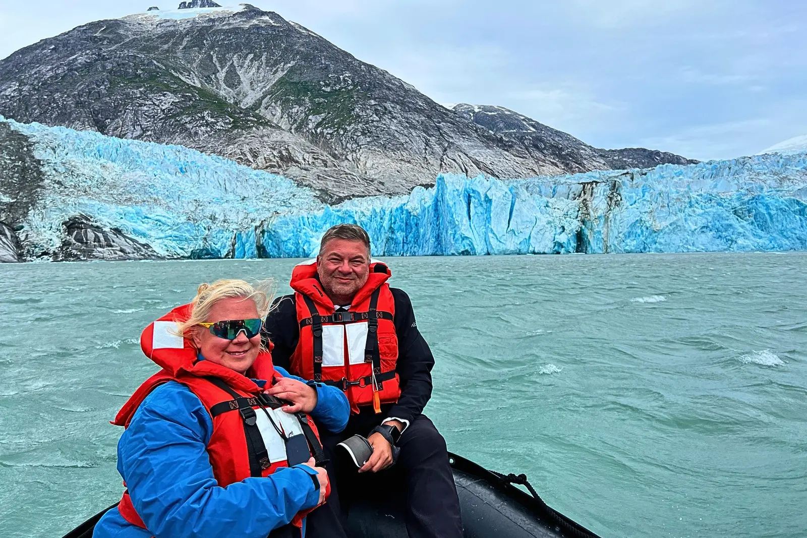 Erika and Chris Commisso in orange life jackets in a Zodiac in front of the blue ice face of Dawes Glacier.