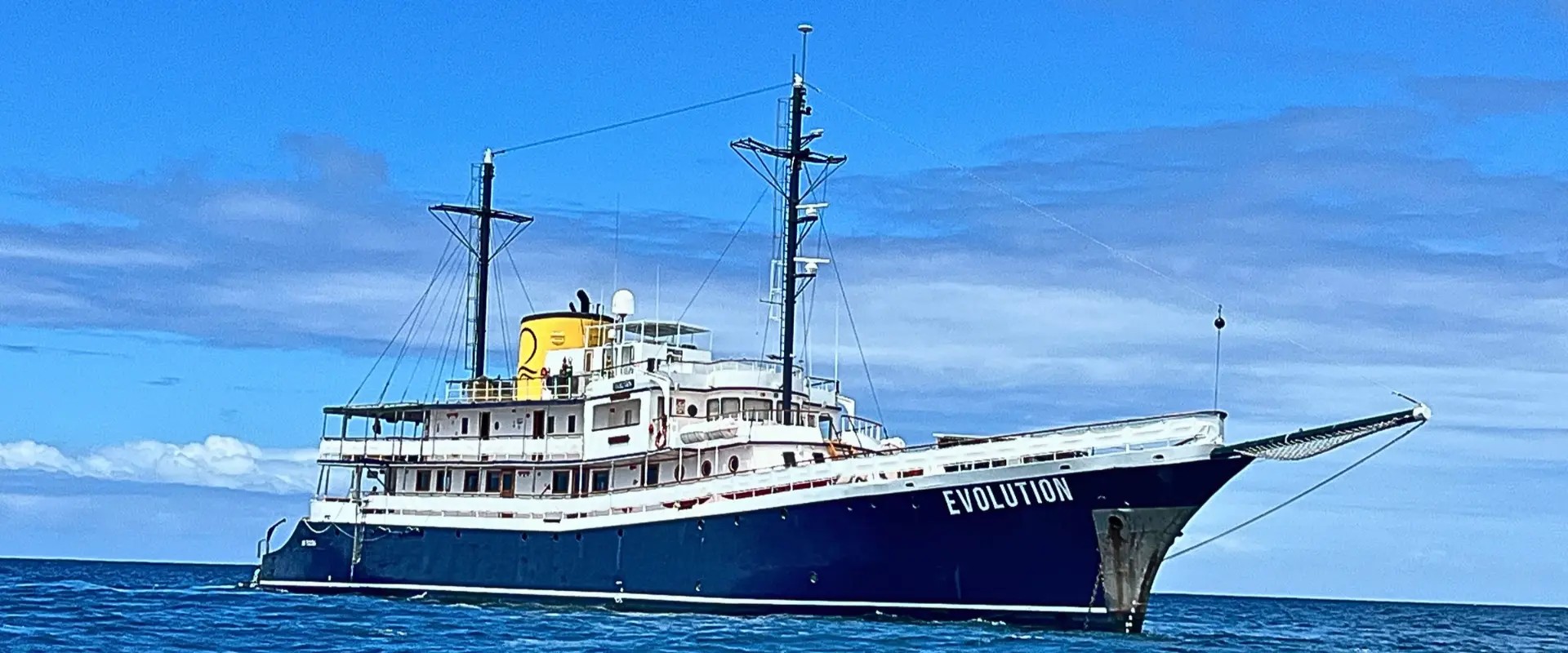 Quasar Expeditions' Evolution at anchor in the Galápagos, with her dark blue hull and yellow funnel under a blue sky.
