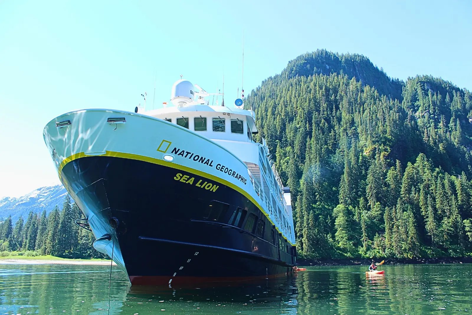 National Geographic Sea Lion anchored in a forested cove, seen from water level, with a kayaker paddling alongside.
