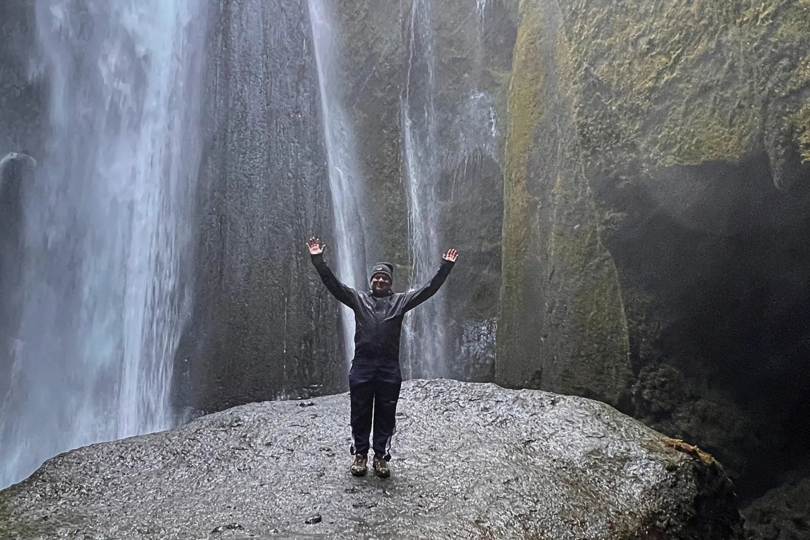 Chris Commisso with arms raised on a rock inside the narrow canyon of Gljúfrabúi, with the waterfall pouring down beside him.