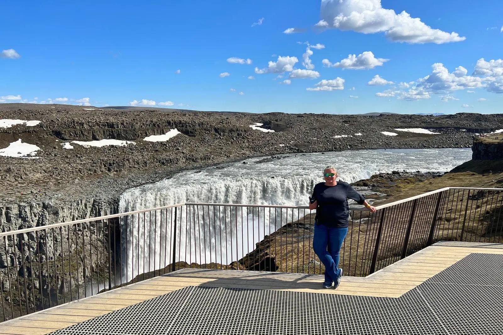 Erika Commisso on a viewing platform in front of Dettifoss, with snow patches on the plateau and blue sky.