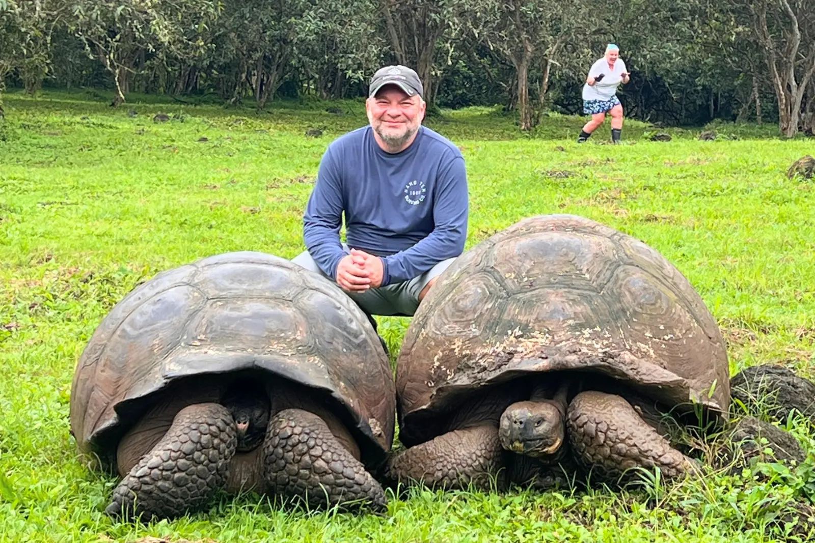 Chris Commisso crouching between two giant tortoises on green grass at El Chato, with Erika in the background.