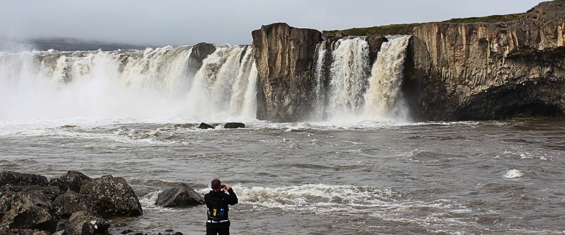 Goðafoss waterfall pouring over dark basalt cliffs in north Iceland, with a visitor photographing it from the rocks.