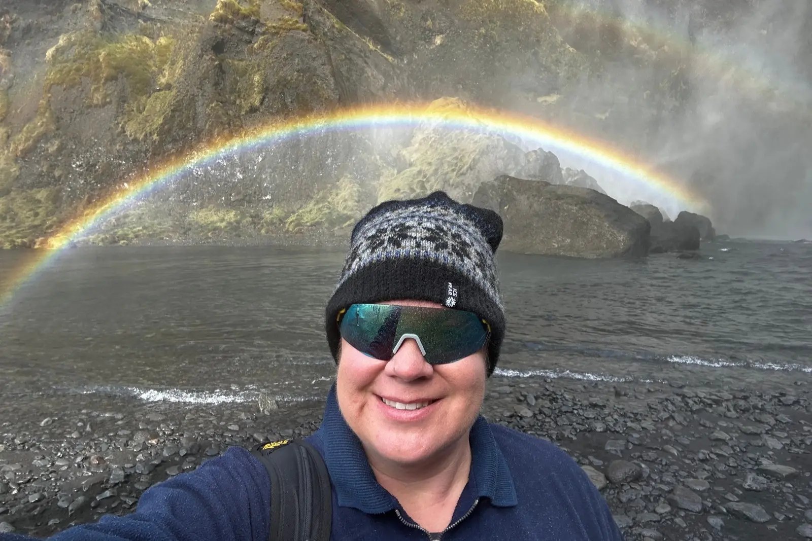 Erika Commisso in a patterned toque and sunglasses at Seljalandsfoss, with a rainbow arching over the waterfall pool behind her.