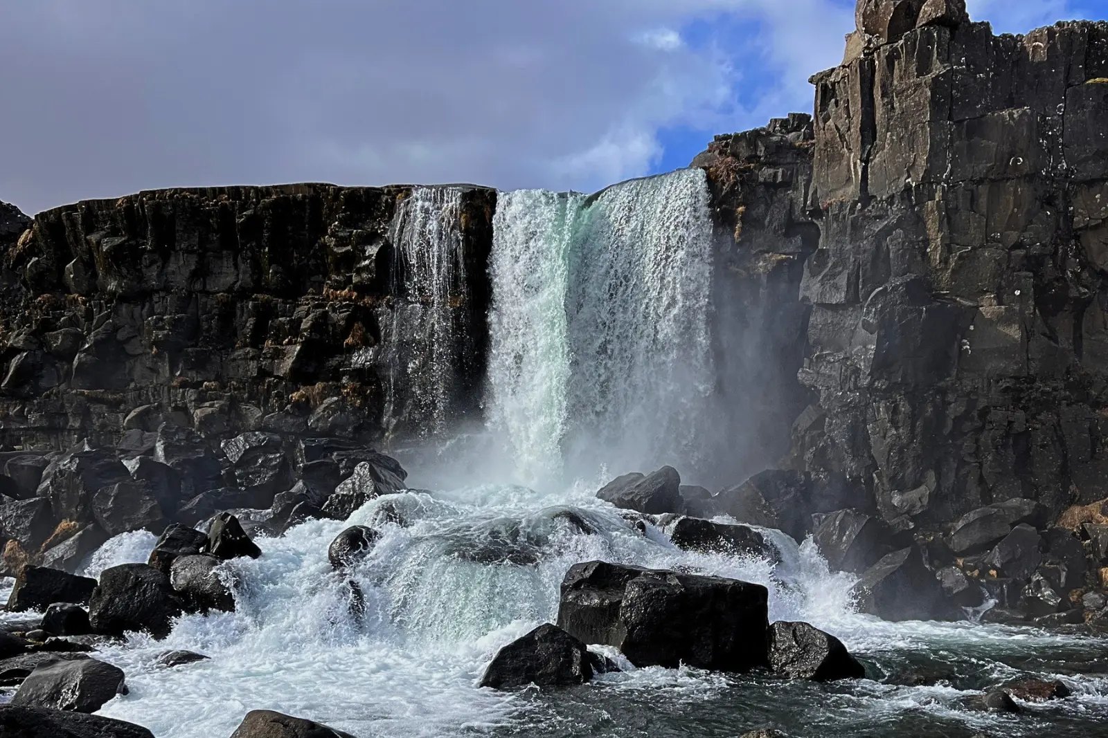 Öxarárfoss pouring through a gap in dark basalt cliffs into a boulder-strewn river in Þingvellir National Park.