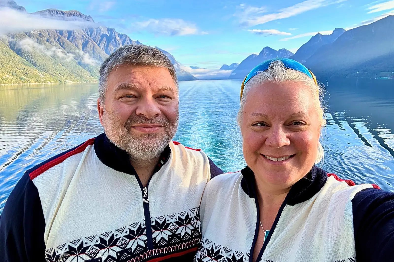 Chris and Erika Commisso in matching Norwegian sweaters, with a fjord, mountains and the ship's wake behind them.
