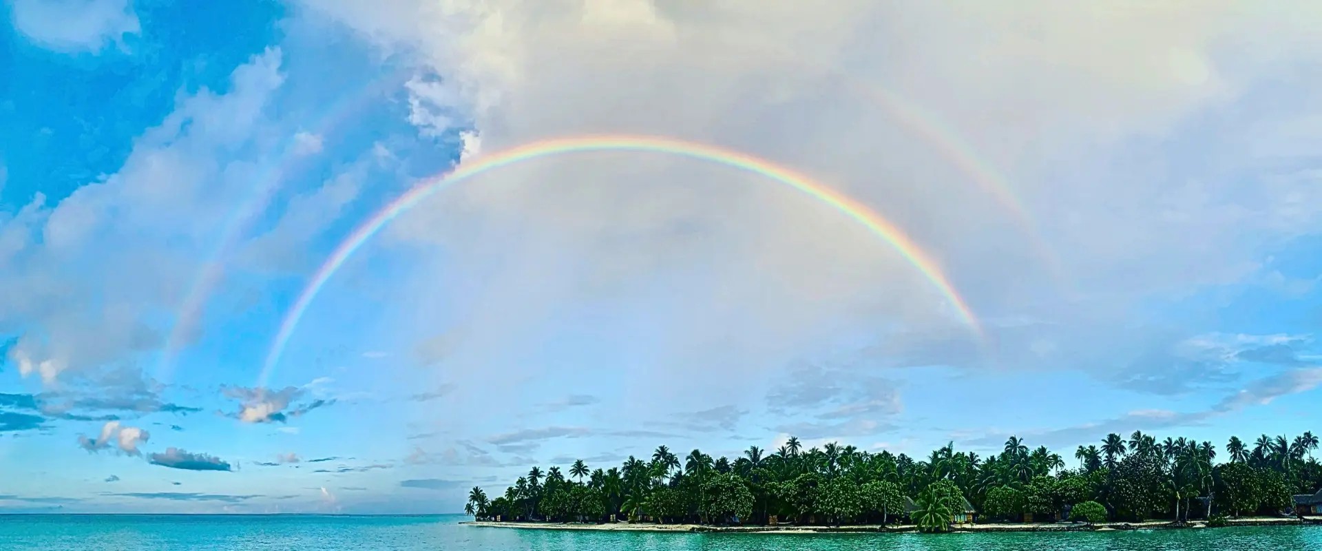 A double rainbow arching over a palm-covered island and turquoise lagoon in French Polynesia.