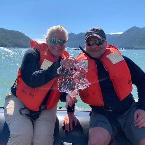 Chris and Erika Commisso in a Zodiac wearing orange life jackets, holding a piece of clear glacial ice in an Alaskan bay.