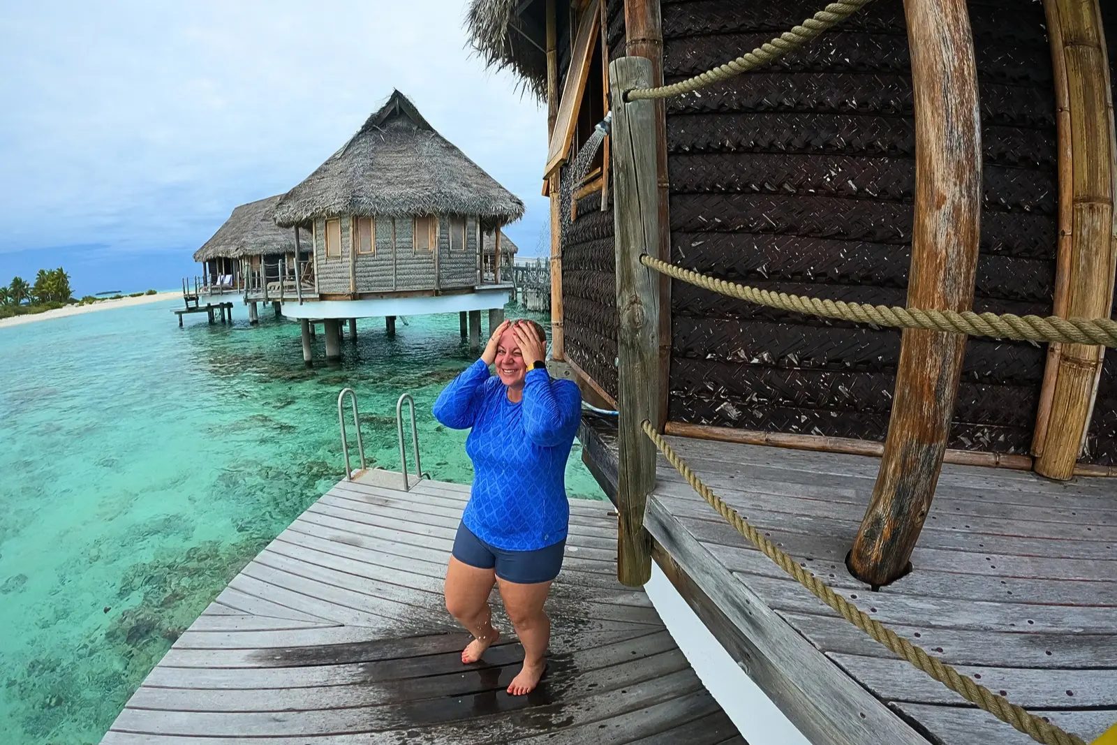 Erika Commisso under the outdoor shower on an overwater bungalow deck at Le Tikehau, with turquoise water and thatched bungalows behind.
