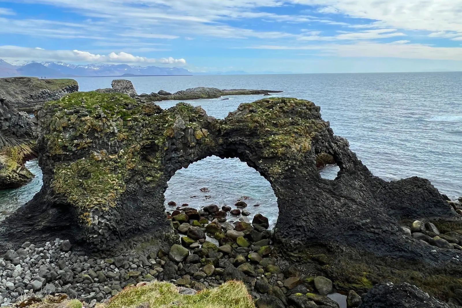 A moss-covered basalt arch over a pebble beach on the Snæfellsnes coast, with the sea and mountains behind.