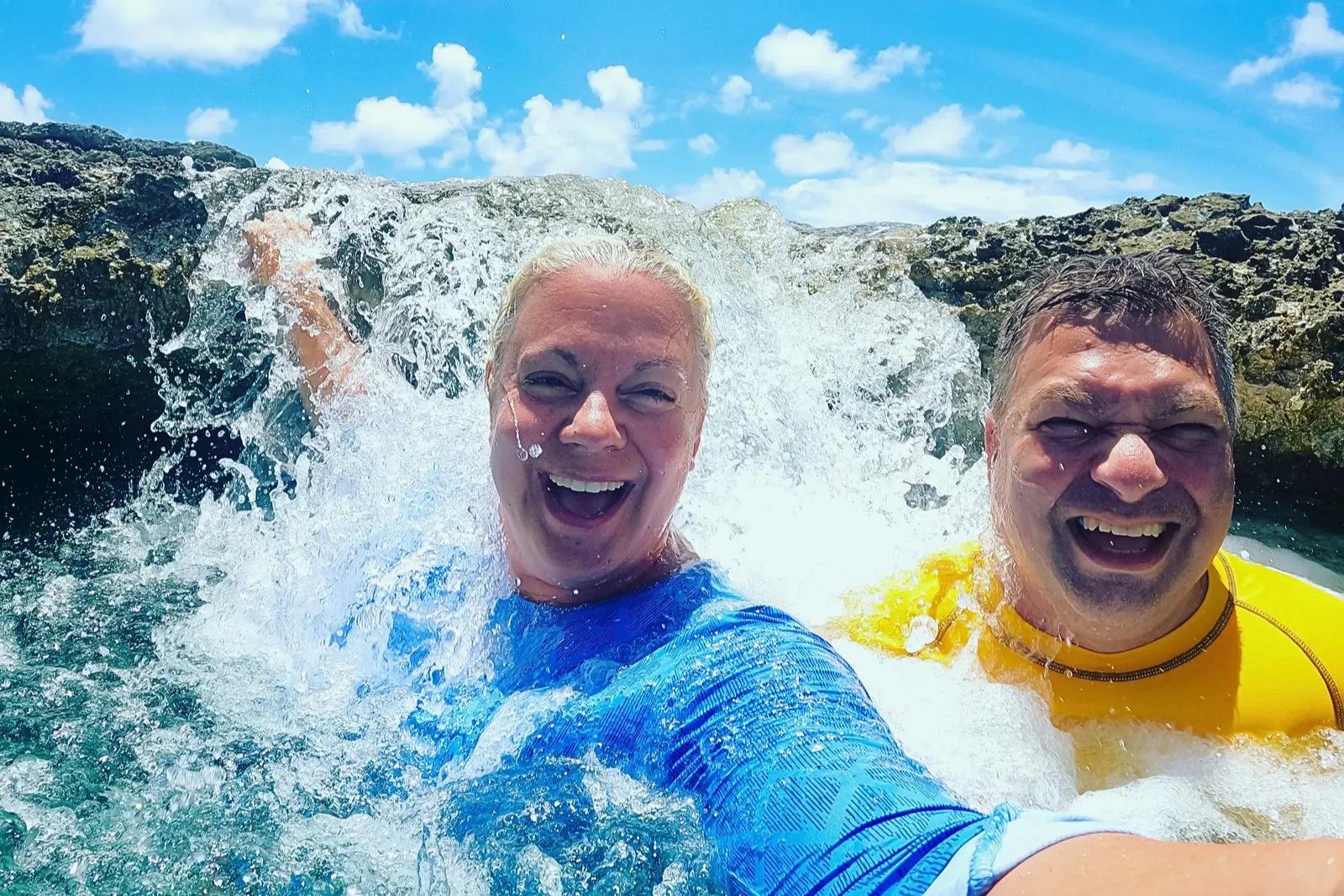 Erika and Chris Commisso laughing as a wave splashes over them against dark rock in Bora Bora.