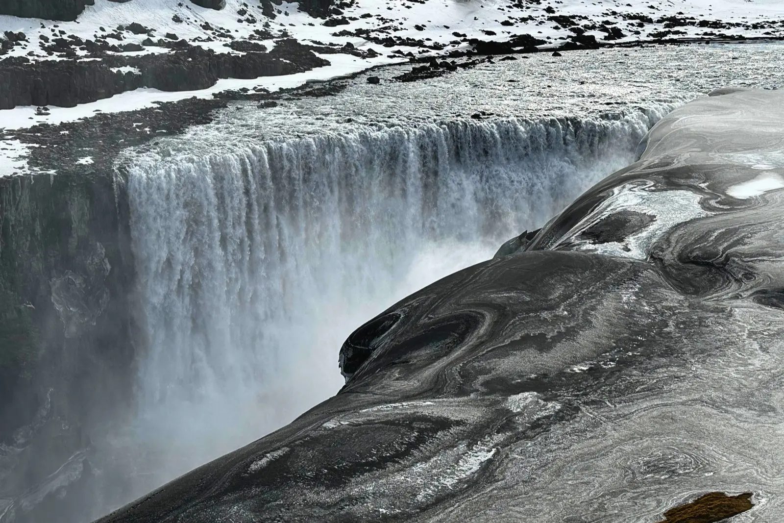 Dettifoss seen from above, a wide curtain of water, with snow and ice on the rock beside it.