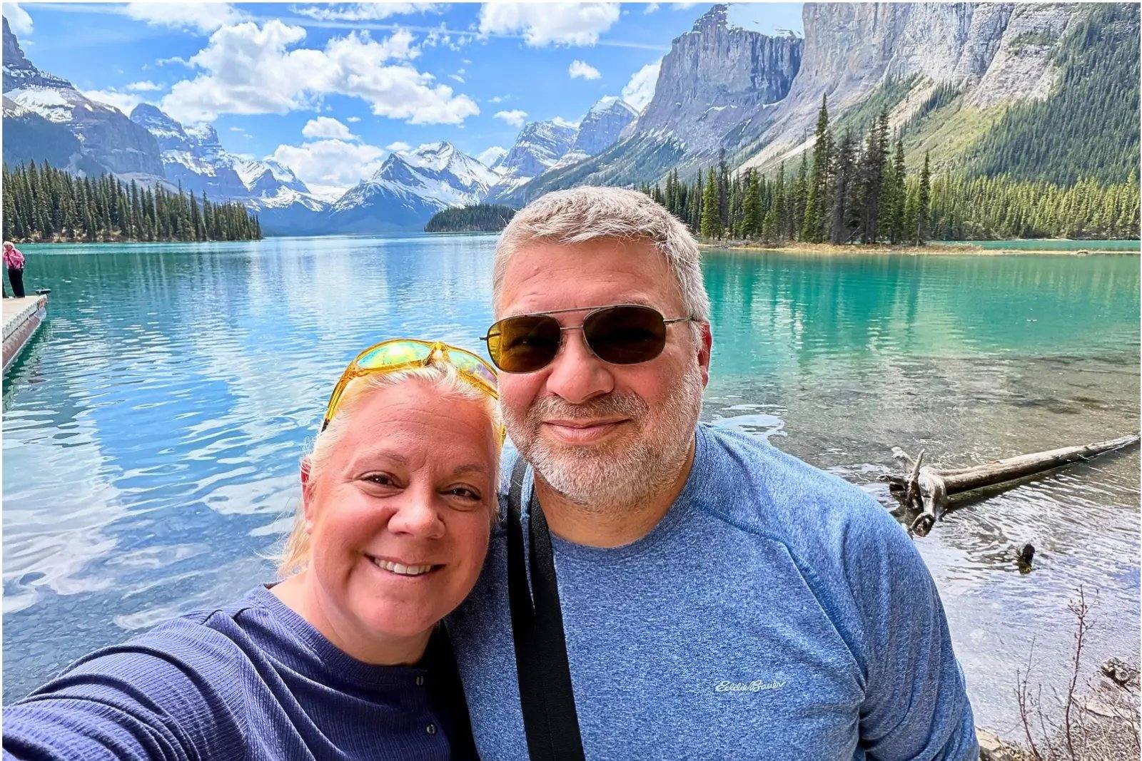 Erika and Chris Commisso taking a selfie beside Maligne Lake, with the boat dock, forest and mountains behind them.