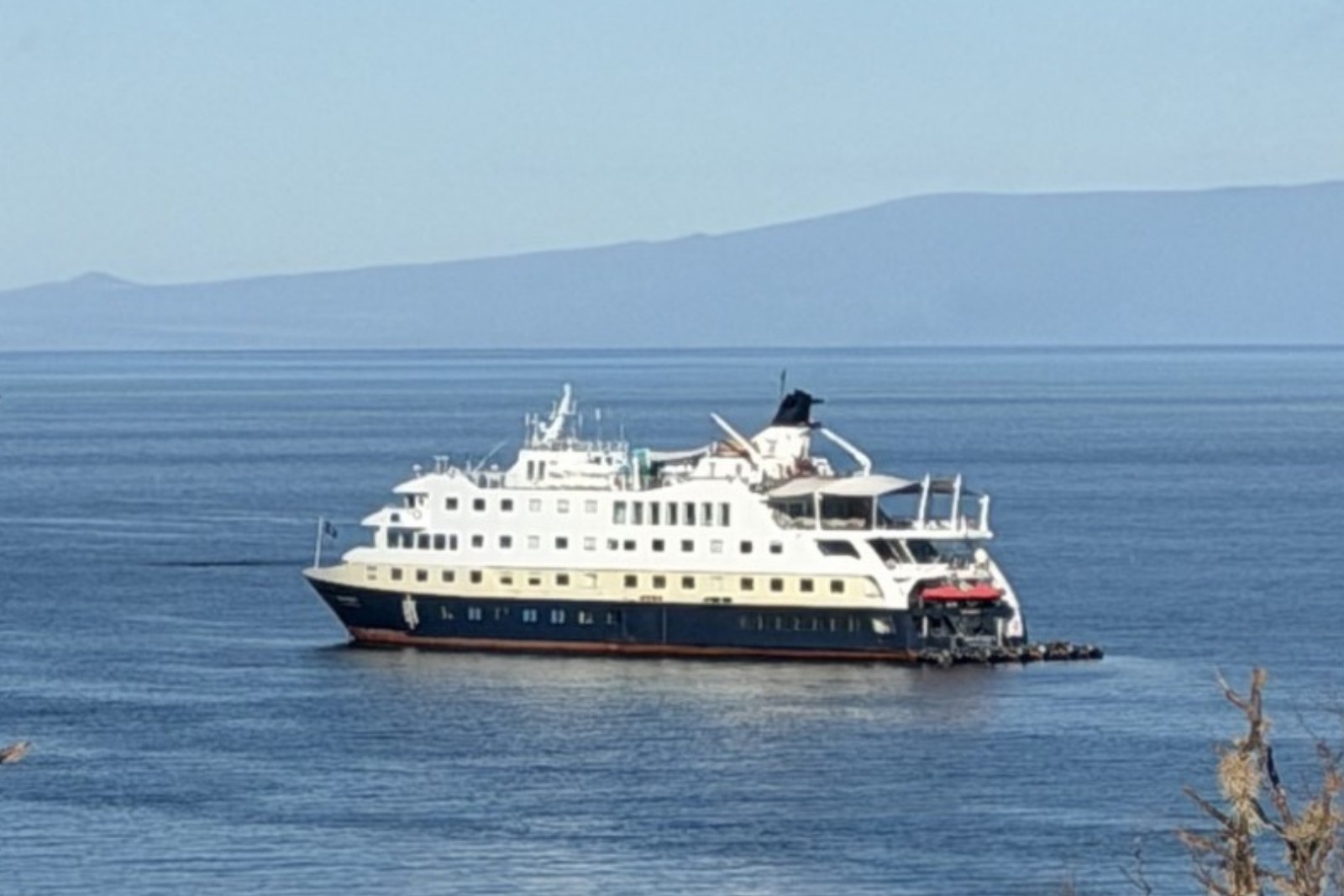HX's Santa Cruz II at anchor in calm blue water in the Galápagos, with a long volcanic island behind her.