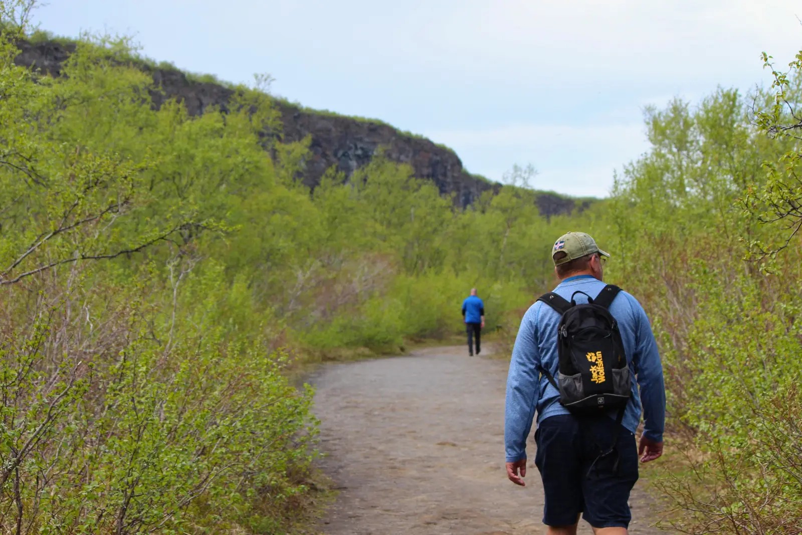 Chris Commisso walking a gravel trail through green shrubs below a dark cliff in north Iceland.