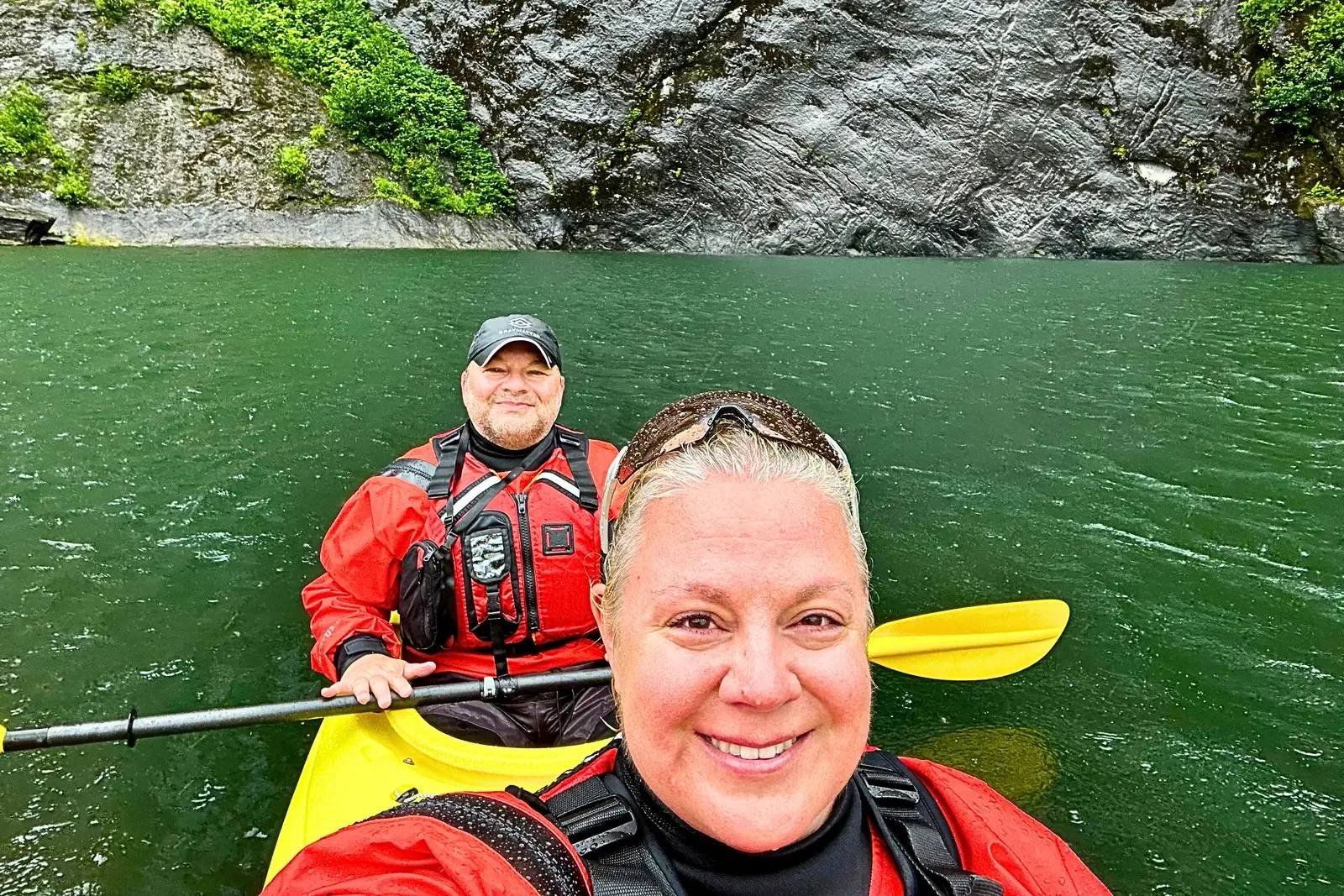 Erika and Chris Commisso in a yellow tandem kayak beside a dark rock wall in the rain in Misty Fjords.