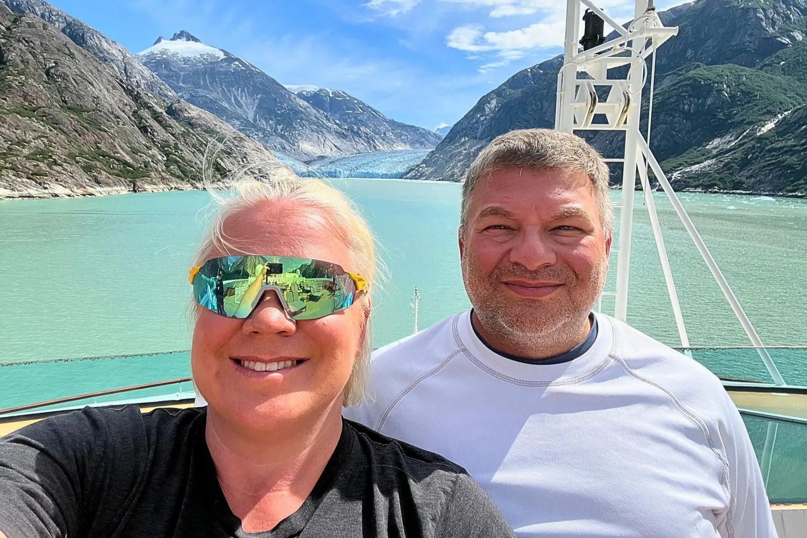 Erika and Chris Commisso taking a selfie on the top deck of Seabourn Odyssey, with turquoise water and Dawes Glacier at the end of Endicott Arm.