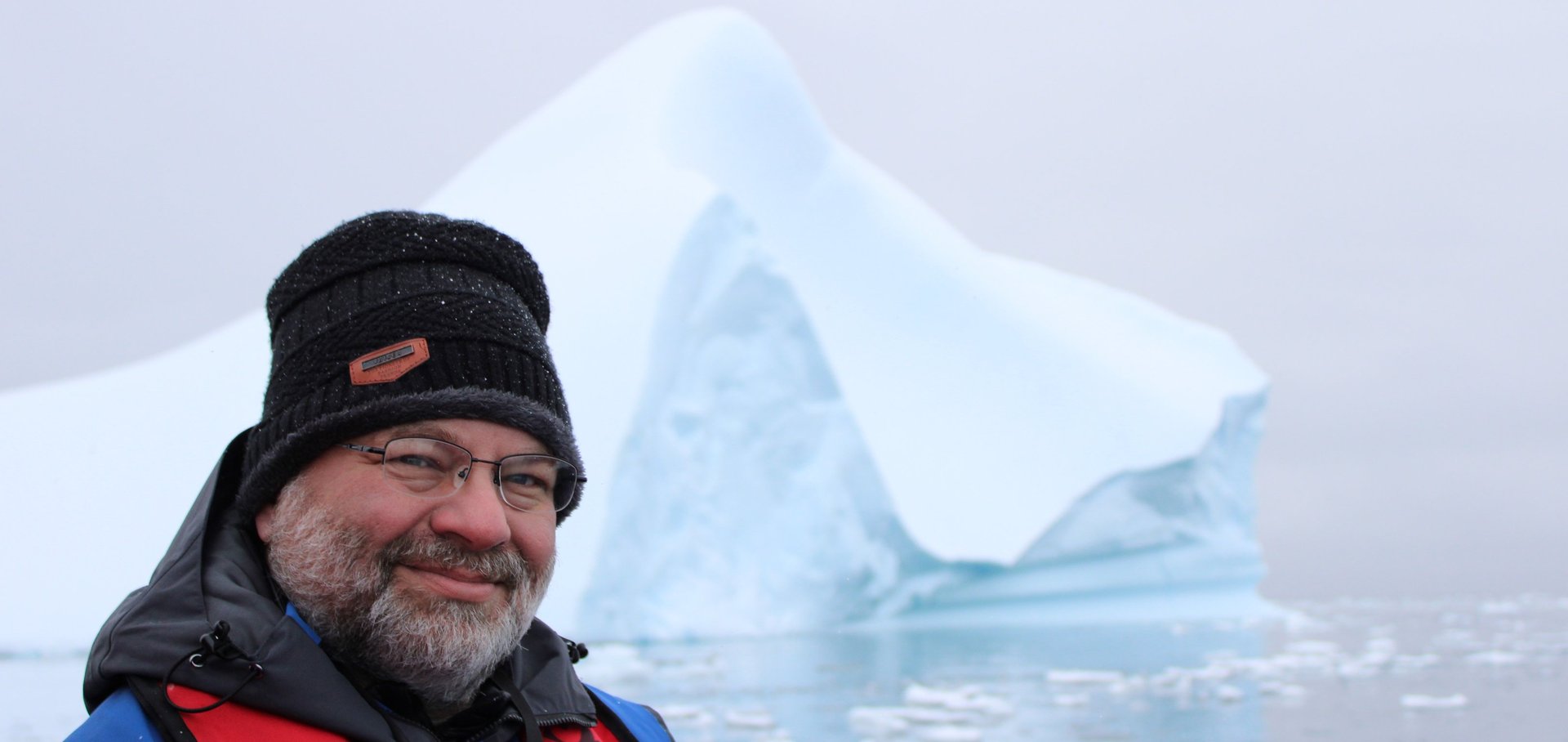 An expedition traveler in a knit hat and parka smiling before a sculpted Antarctic iceberg.