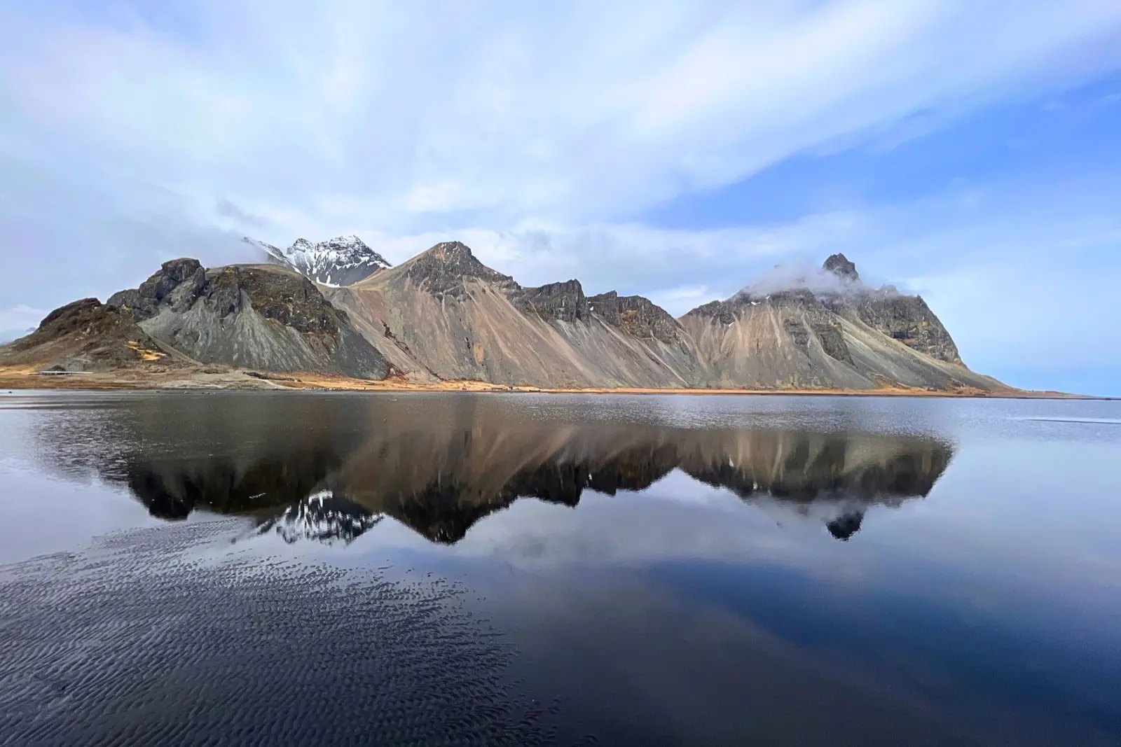 The jagged peaks of Vestrahorn reflected in still water on the black sand shore near the Viking Village.
