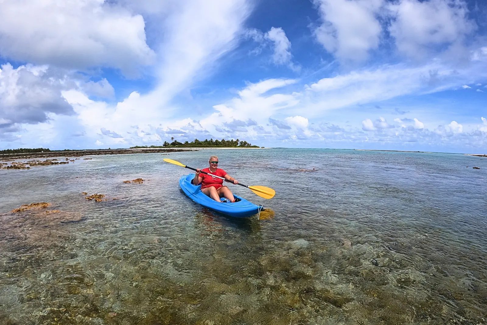 Erika Commisso paddling a blue kayak over a shallow reef on Tikehau, with a palm-covered motu behind her.