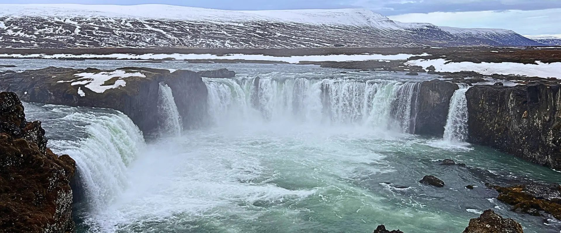 Goðafoss in April, pouring in a horseshoe over dark rock, with snow on the banks and snow-covered hills behind.