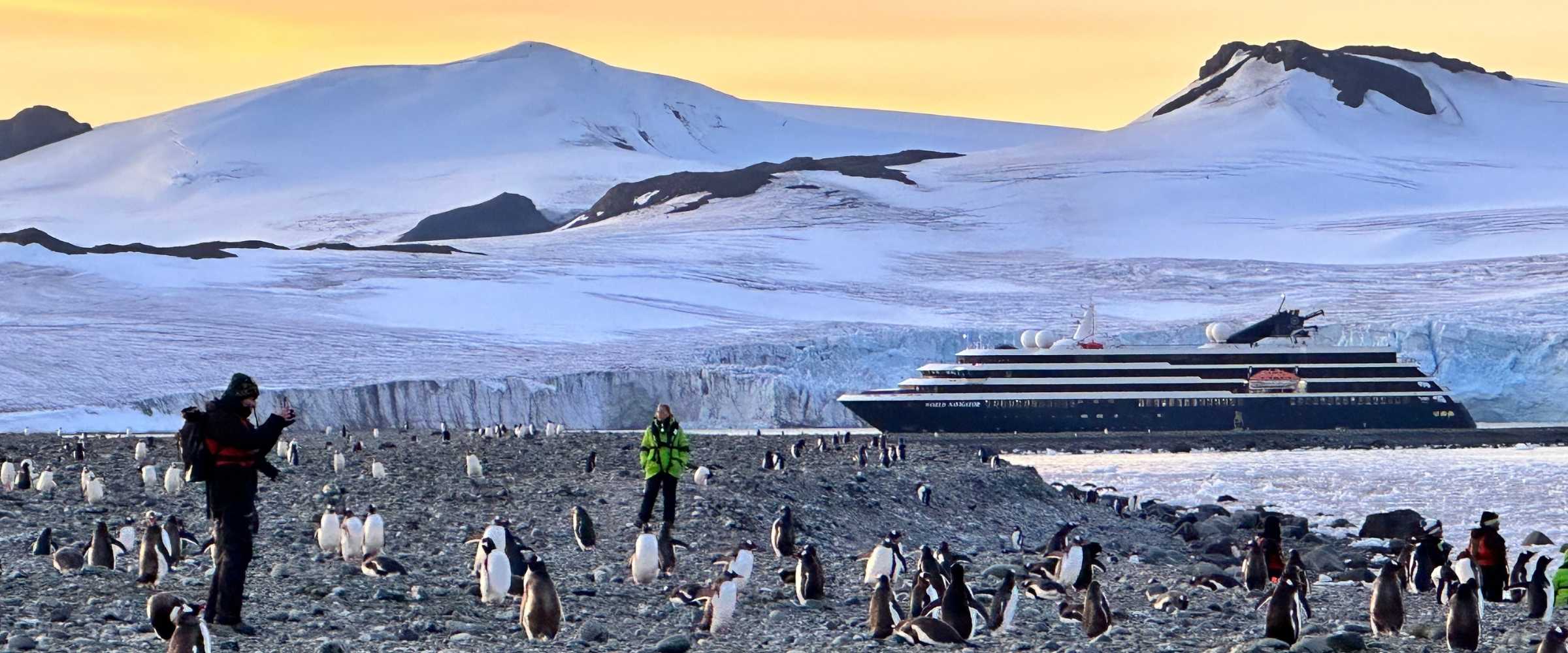 Gentoo penguins and the World Navigator in front of a glacier and snow-covered Antarctic mountains.