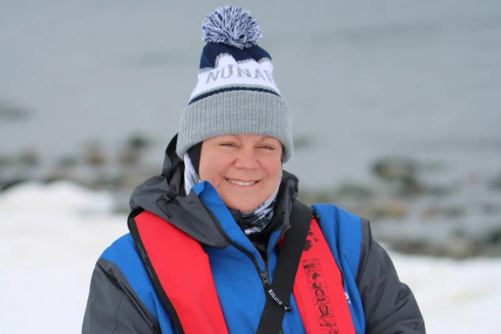 Erika Commisso smiling in a toque, blue Aurora parka and red life jacket, with snow and dark water behind her.