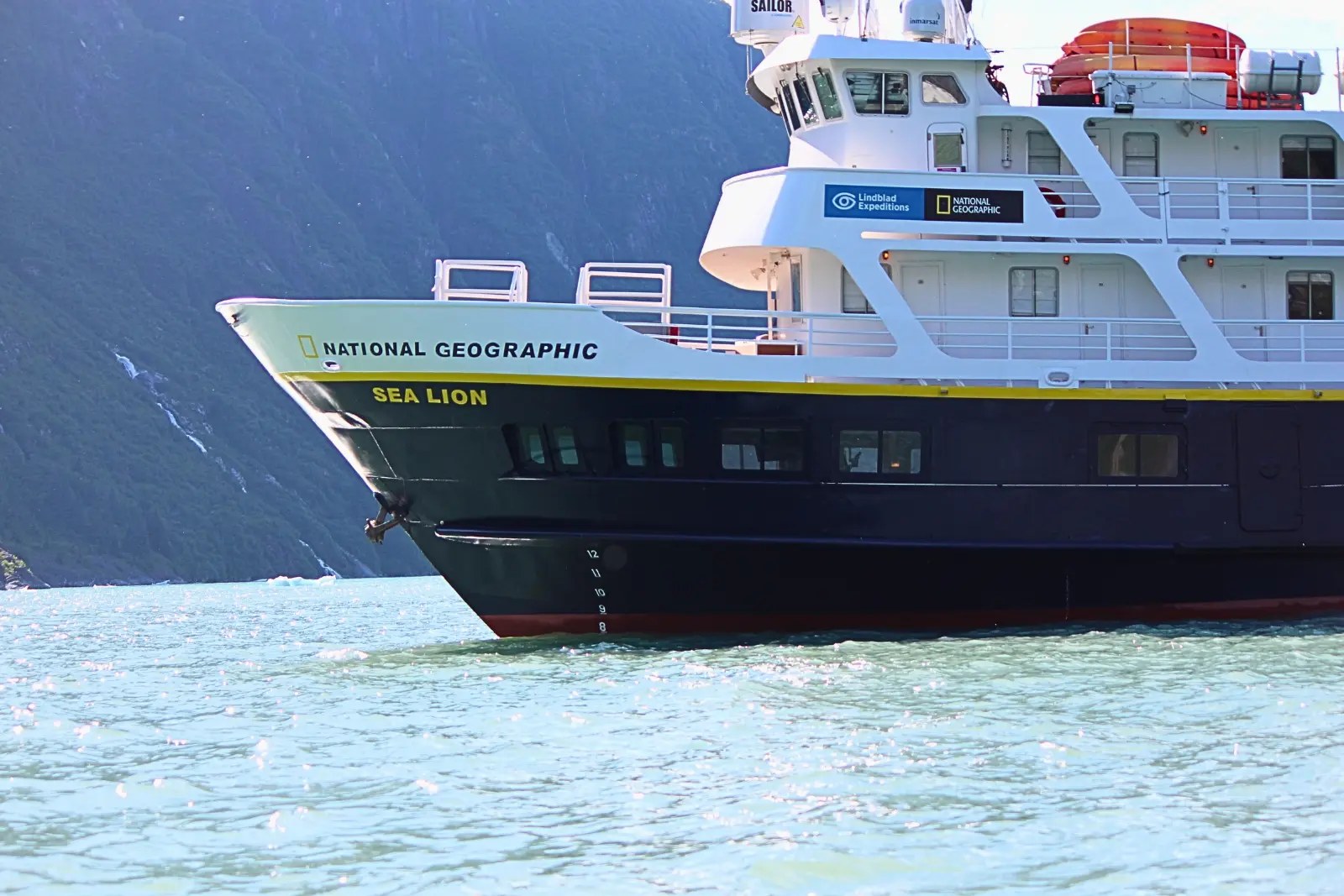 The bow of National Geographic Sea Lion, with Lindblad Expeditions and National Geographic logos, below a steep fjord wall.