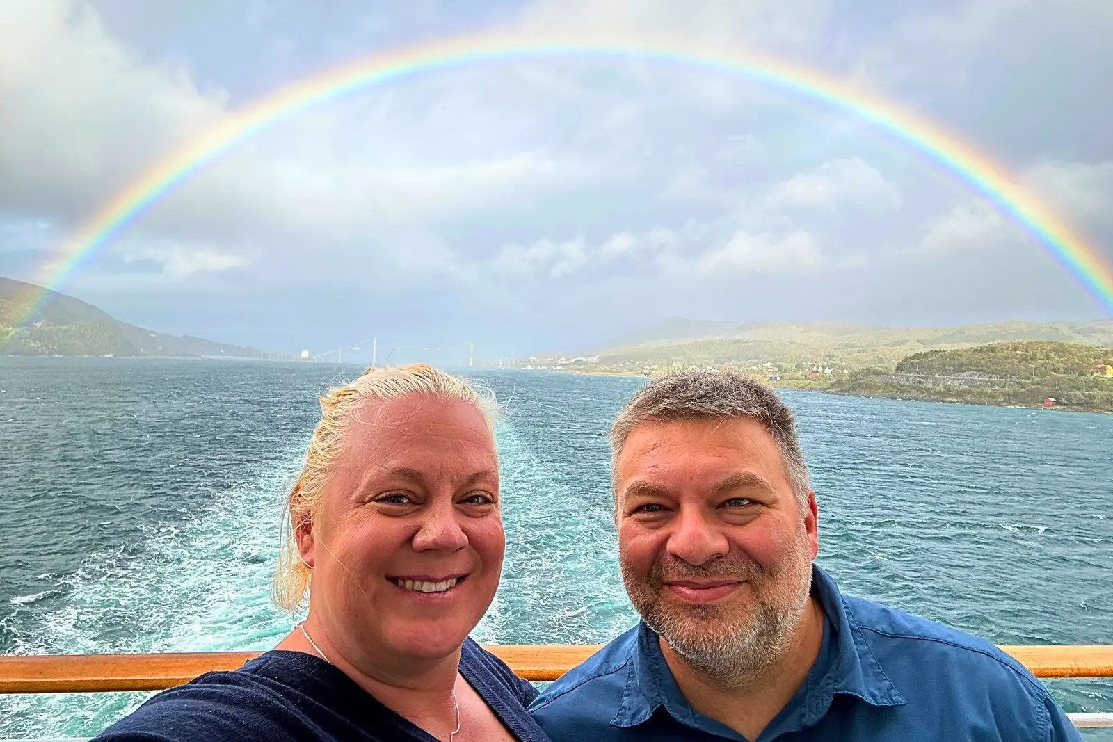 Erika and Chris Commisso taking a selfie on deck with a full rainbow over the water behind them.