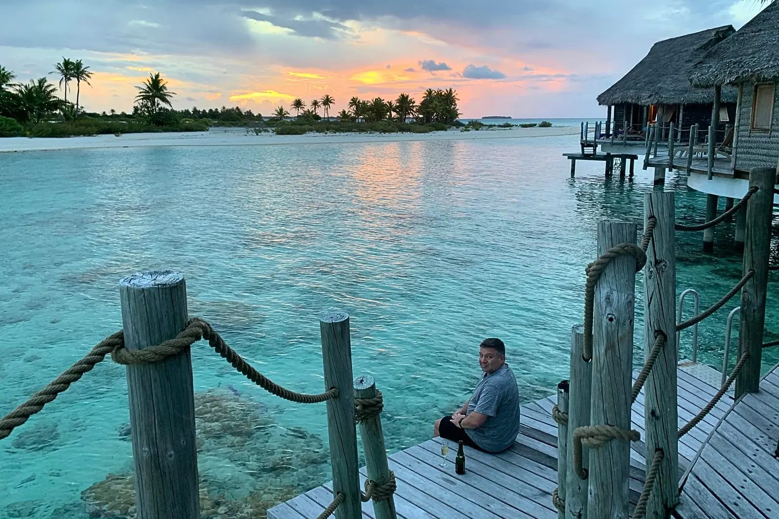 Chris Commisso sitting on a bungalow dock at sunset on Tikehau with a glass and a bottle, palms and a sandbar behind him.