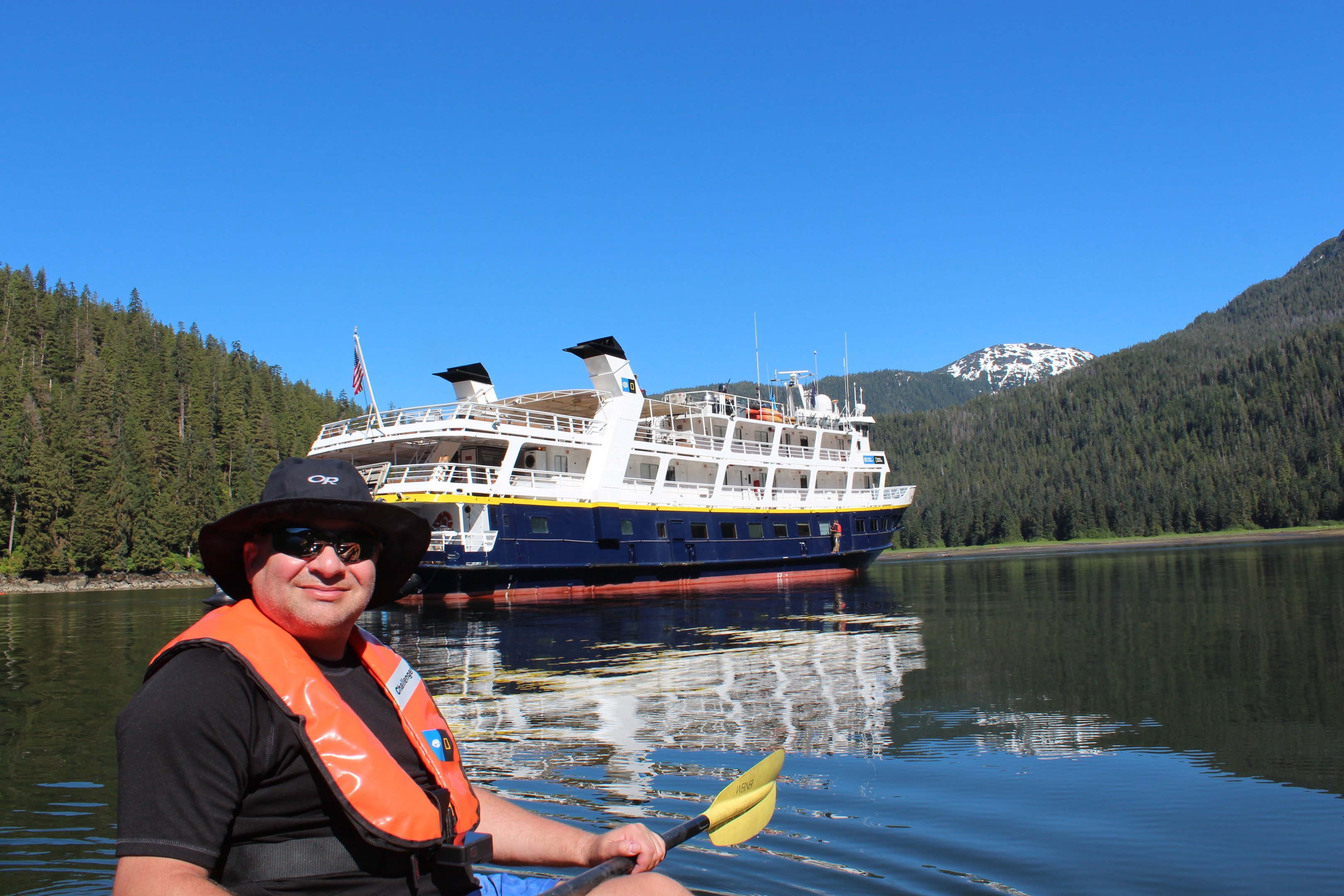 A man kayaking in an orange life vest near a cruise ship in a calm Alaskan bay with mountains.