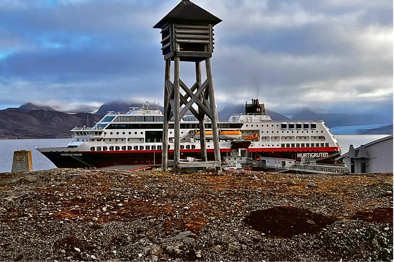 A small weathered wooden tower on gravel, with the Trollfjord docked behind it.