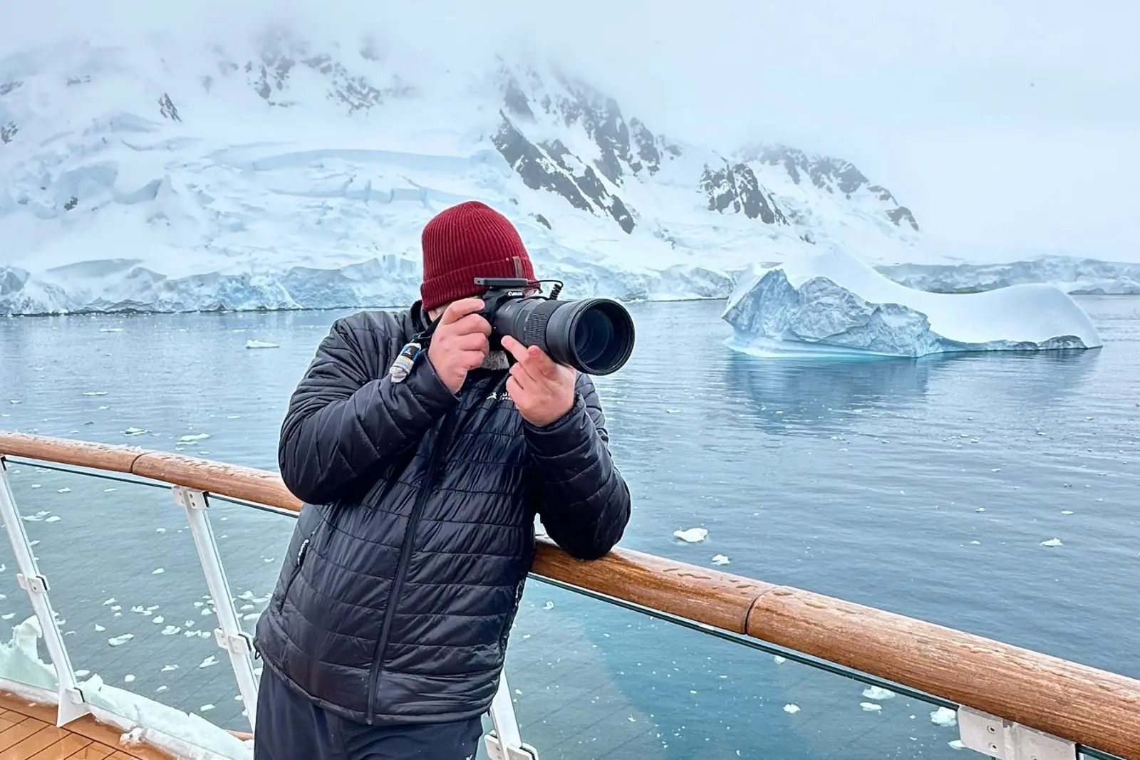 Chris Commisso photographing from the Greg Mortimer's rail with a long lens, with a glacier and iceberg behind him in fog.