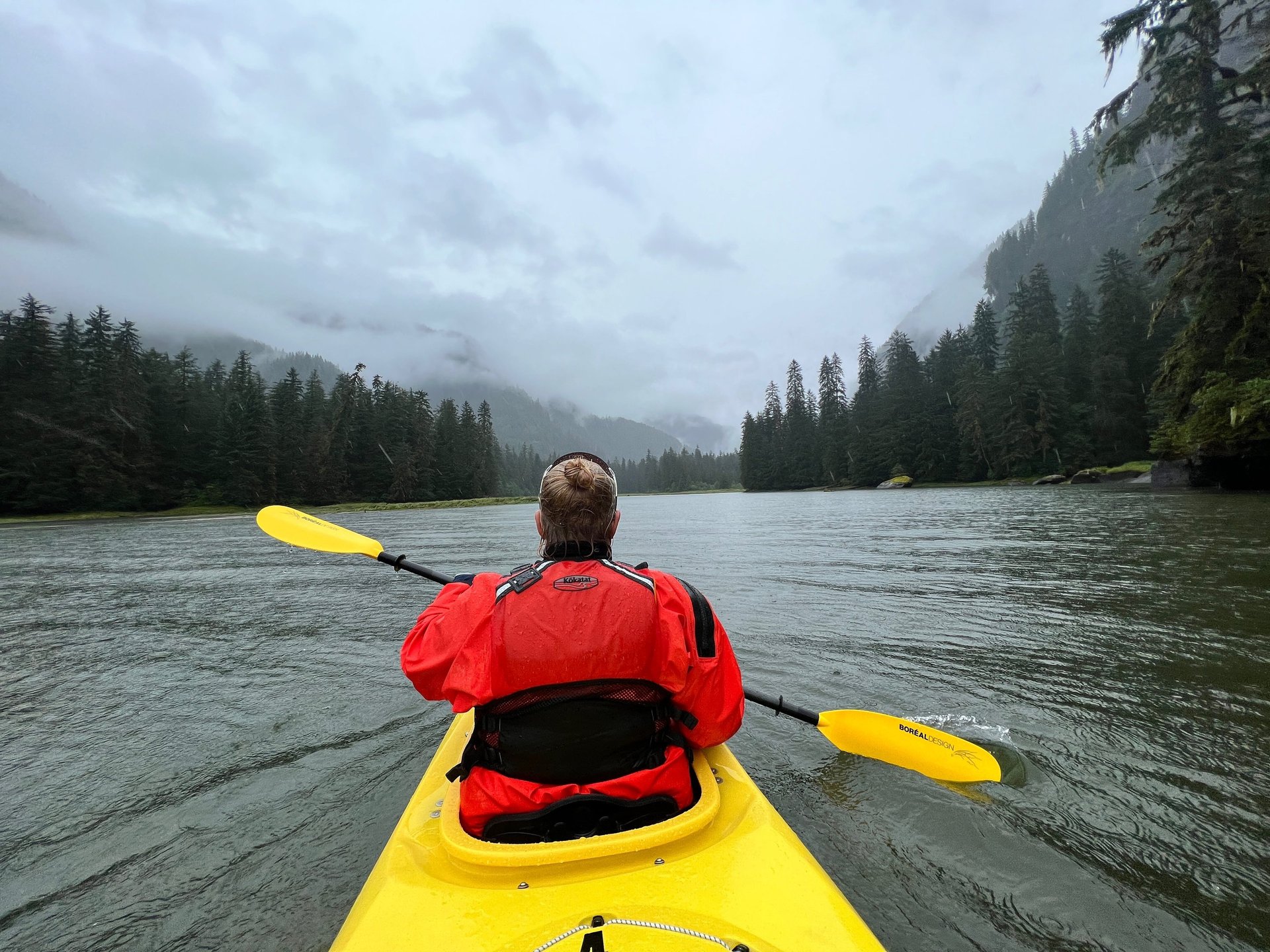 A paddler in a red jacket kayaking a misty fjord lined with spruce forest.