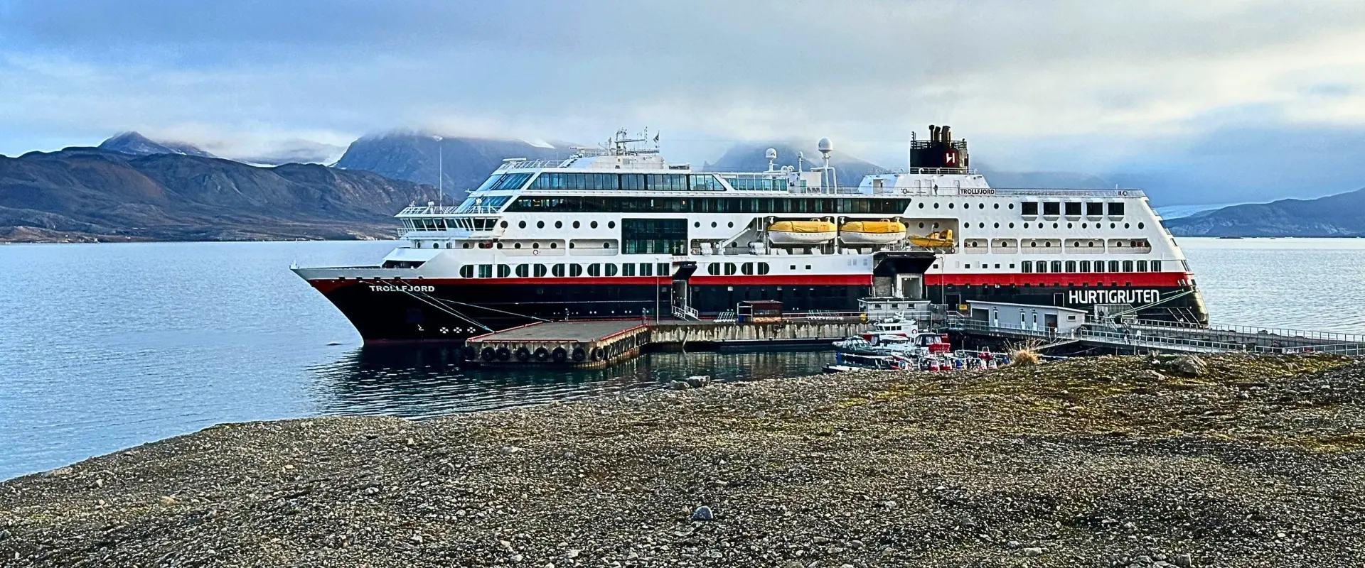 Hurtigruten's Trollfjord docked at a pier in Svalbard, with mountains and a glacier behind her.