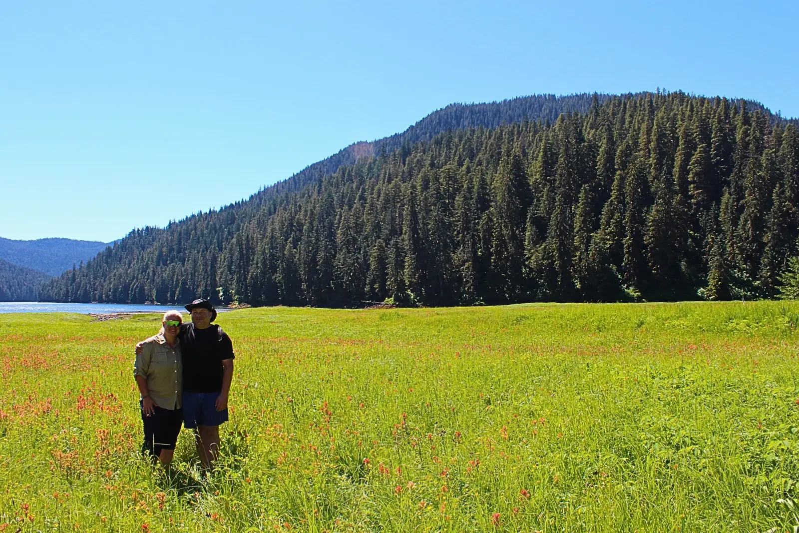 Erika and Chris Commisso standing in a meadow of red wildflowers, with a forested hill and water behind them.