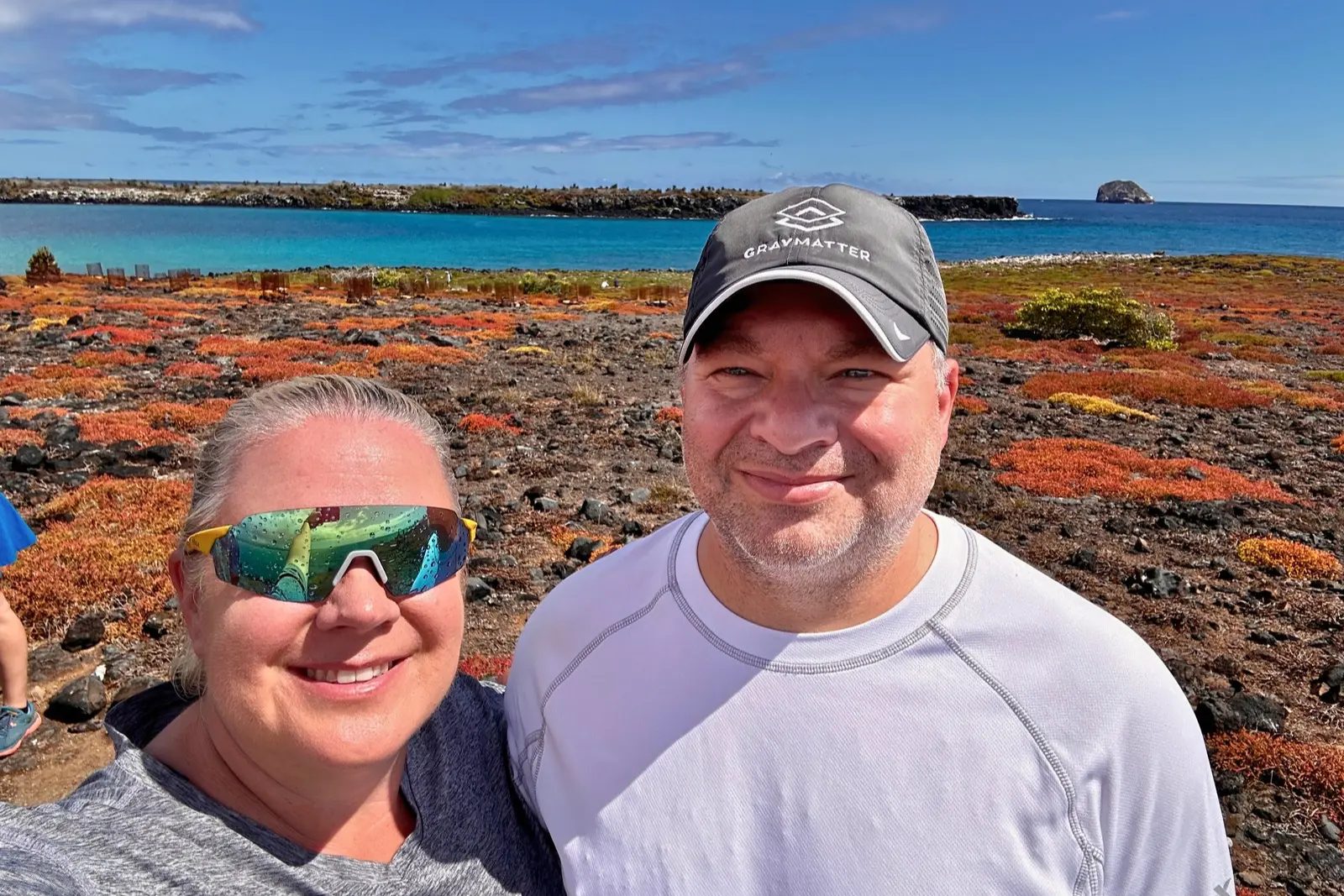 Erika and Chris Commisso taking a selfie on a landscape carpeted in red and orange plants, with turquoise water behind them.