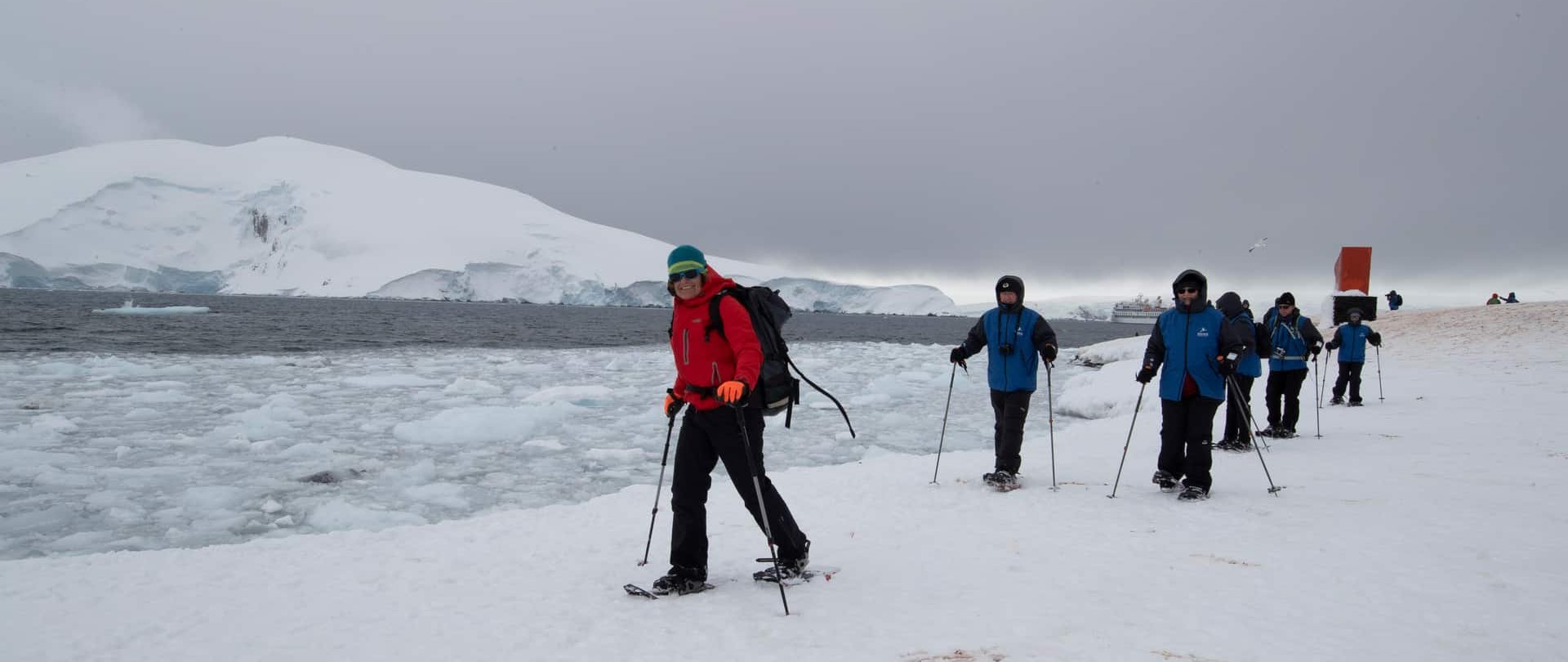 Expedition guests snowshoeing along the Antarctic shoreline with their ship anchored offshore.