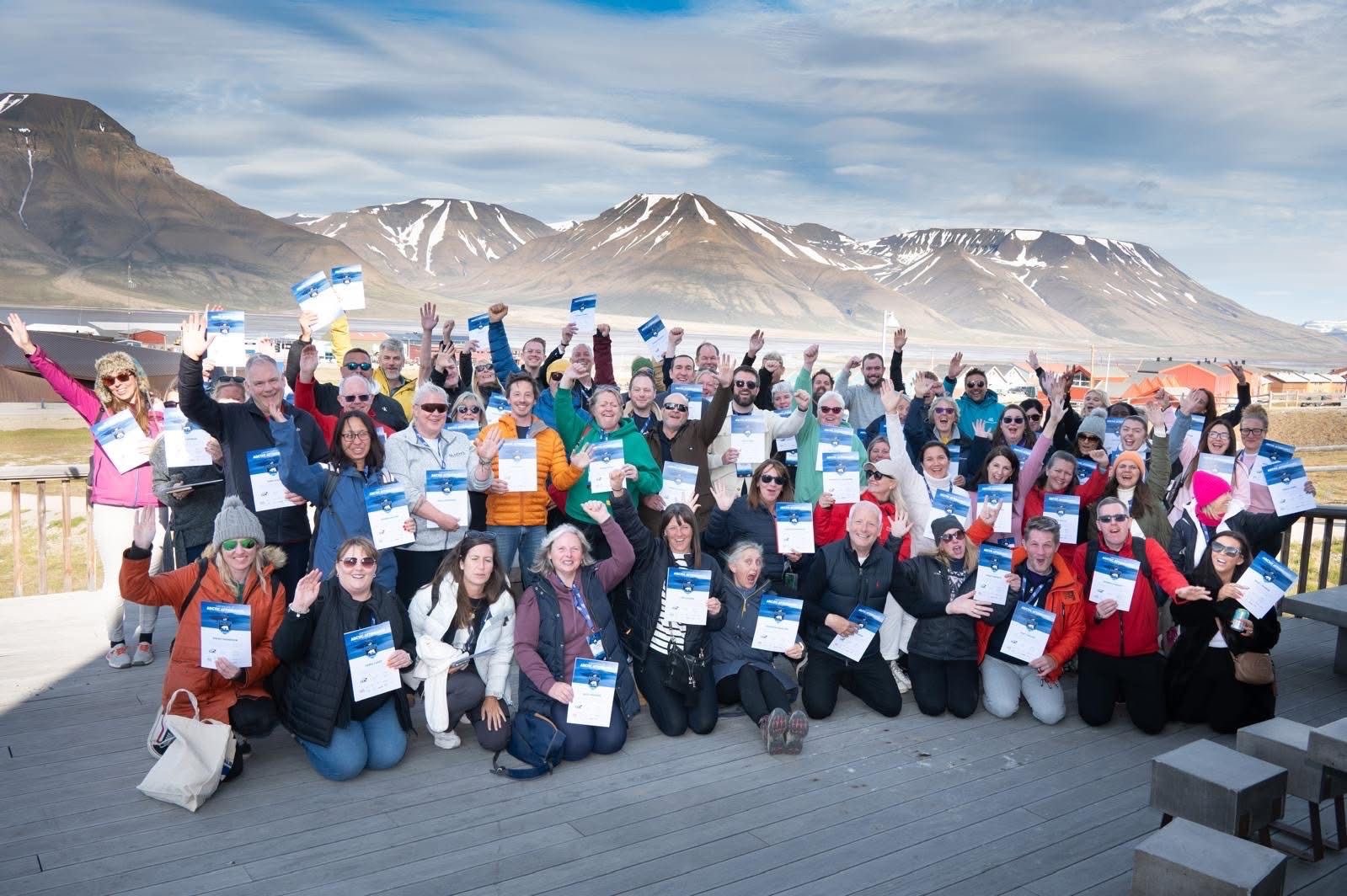A large group of summit participants waving and holding certificates on a deck in Longyearbyen, with mountains behind.
