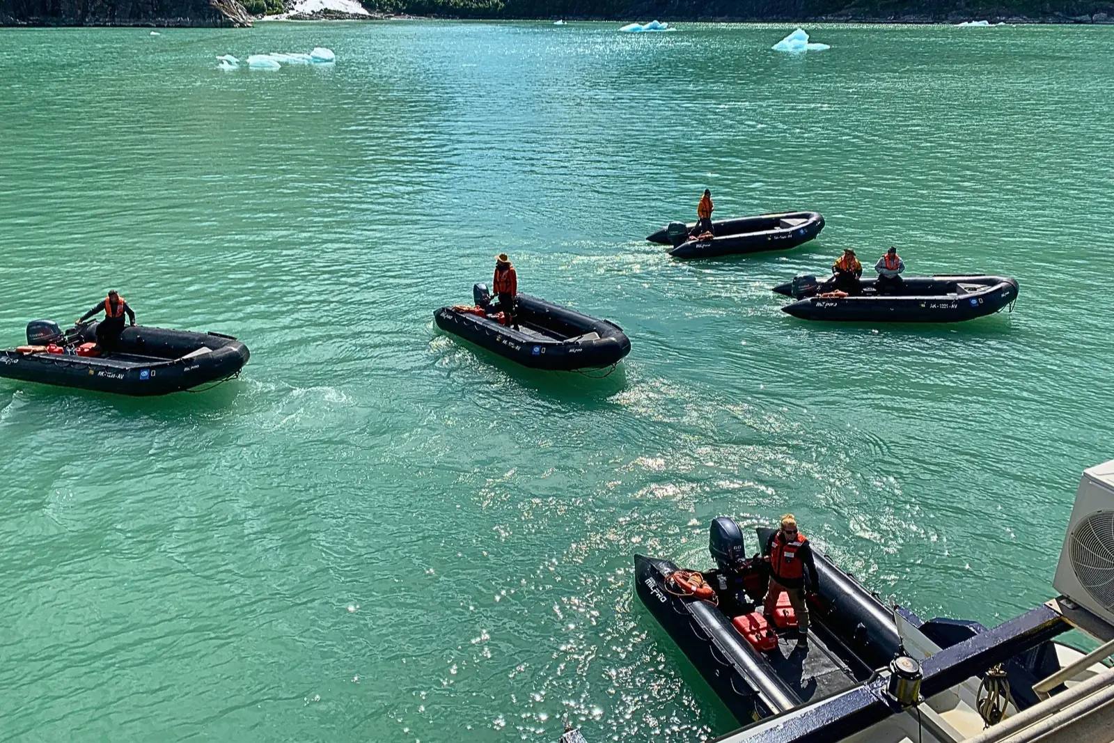 Black Zodiacs with drivers in orange life jackets on turquoise water, seen from the deck of National Geographic Sea Lion.