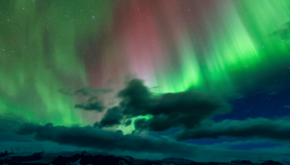 Green and pink Northern Lights dancing above clouds and Arctic ice under a starry sky.