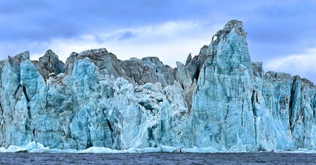 A massive blue glacier ice wall towering over dark ocean water in Svalbard under a cloudy sky.