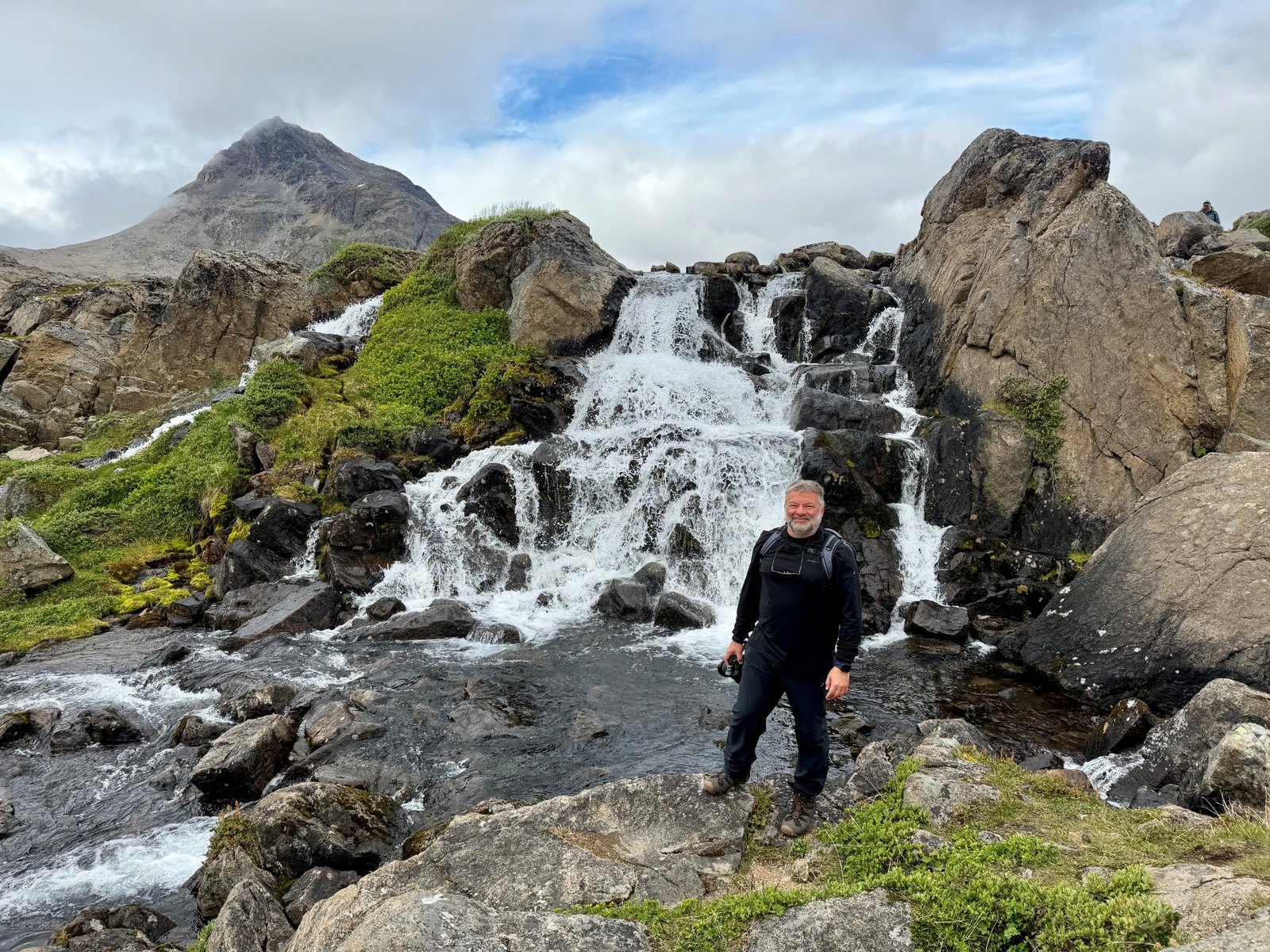 Chris Commisso holding a camera beside a waterfall on a rocky, moss-covered hillside in the Arctic.
